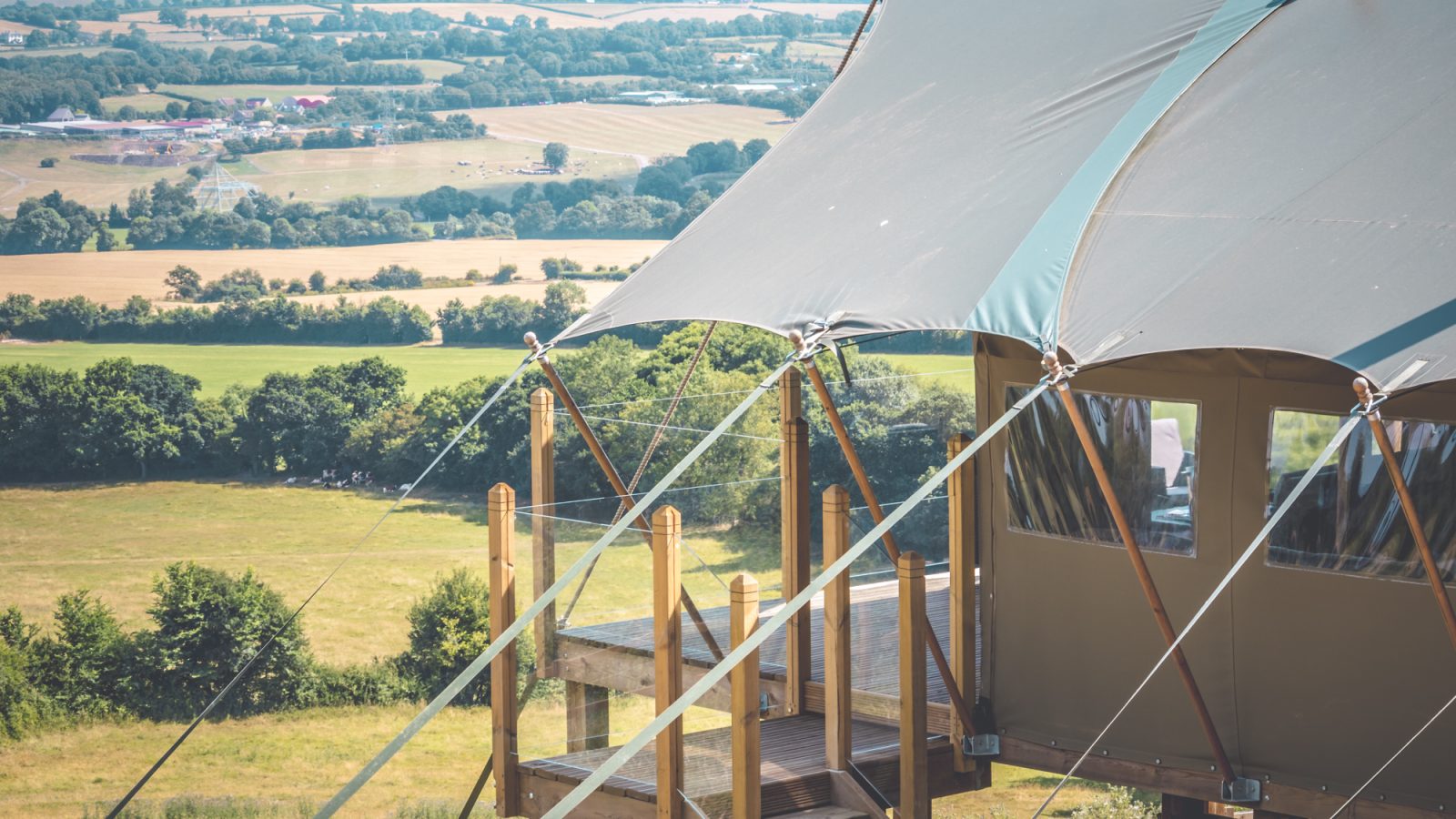 A luxury safari tent with a large canopy roof is set up on a wooden platform overlooking rolling green fields and farmland. The tent has windows, and the structure is secured with ropes. The landscape features a mix of trees and open land under a clear blue sky.