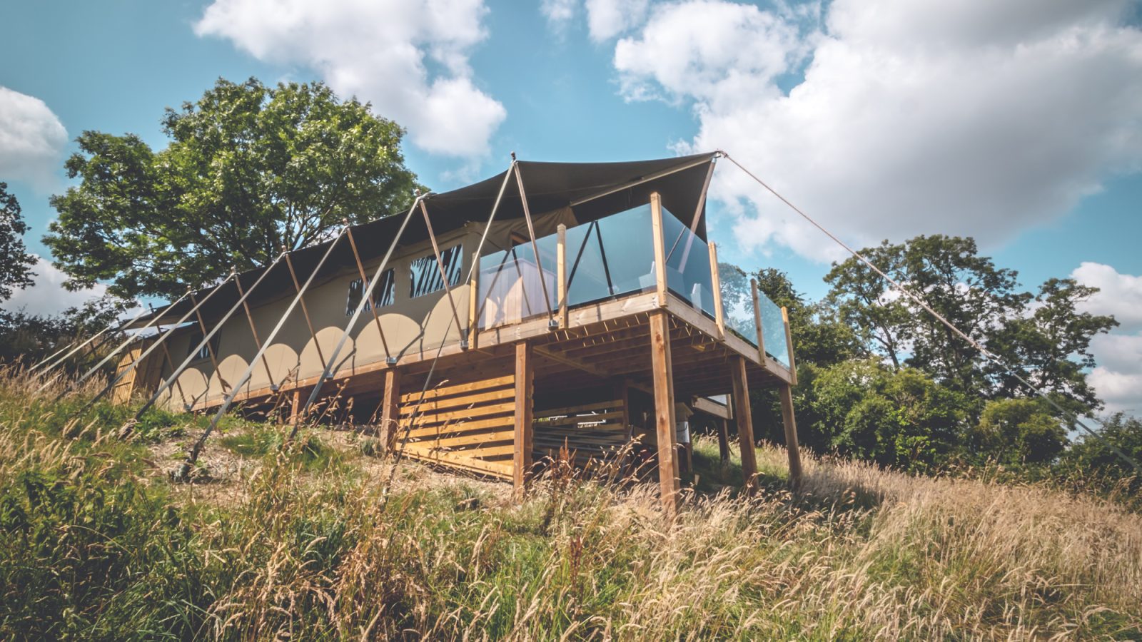 A modern safari tent-like structure with large windows is elevated on stilts in a grassy, hilly area surrounded by trees under a partly cloudy sky. The building features a canopy-style roof and open sides with wooden slats supporting the base.