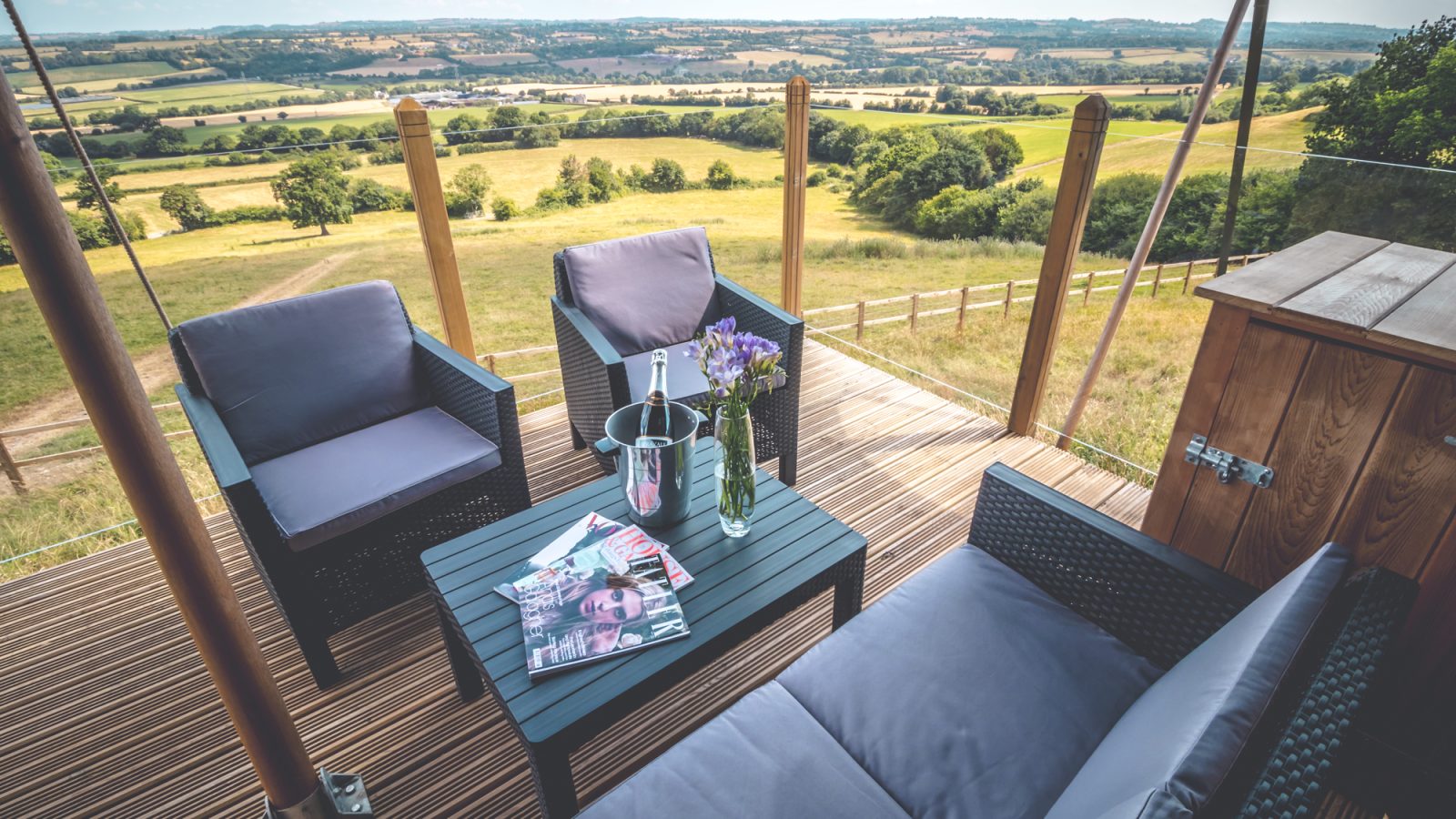 A scenic outdoor patio adjacent to a luxurious safari tent overlooks a vast countryside. The patio features two armchairs and a sofa with gray cushions, a table holding a vase with purple flowers, a wine bottle in an ice bucket, and a stack of magazines.