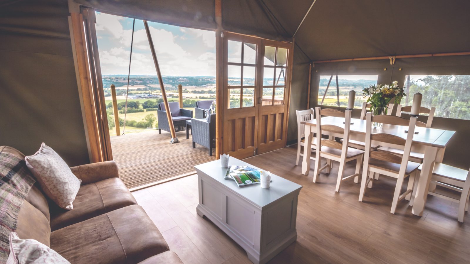 Interior of a spacious Safari tent with a cozy sitting area on the left, featuring a brown sofa and coffee table. On the right is a dining table and chairs. The open door reveals a wooden deck with additional seating and a scenic view of a lush green landscape, perfect for luxury camping.