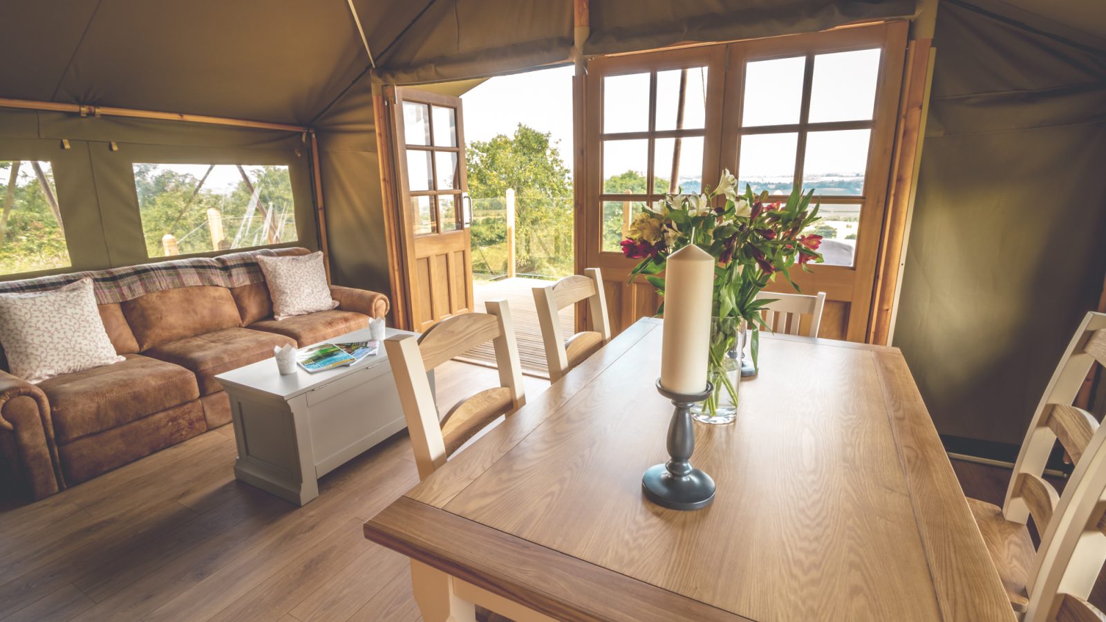 Interior of a cozy safari tent with wooden floors, a comfortable brown sofa, large windows, and a wooden dining table set. The table is decorated with candles and a vase of red and white flowers. The safari tent opens to an outdoor scenic view.