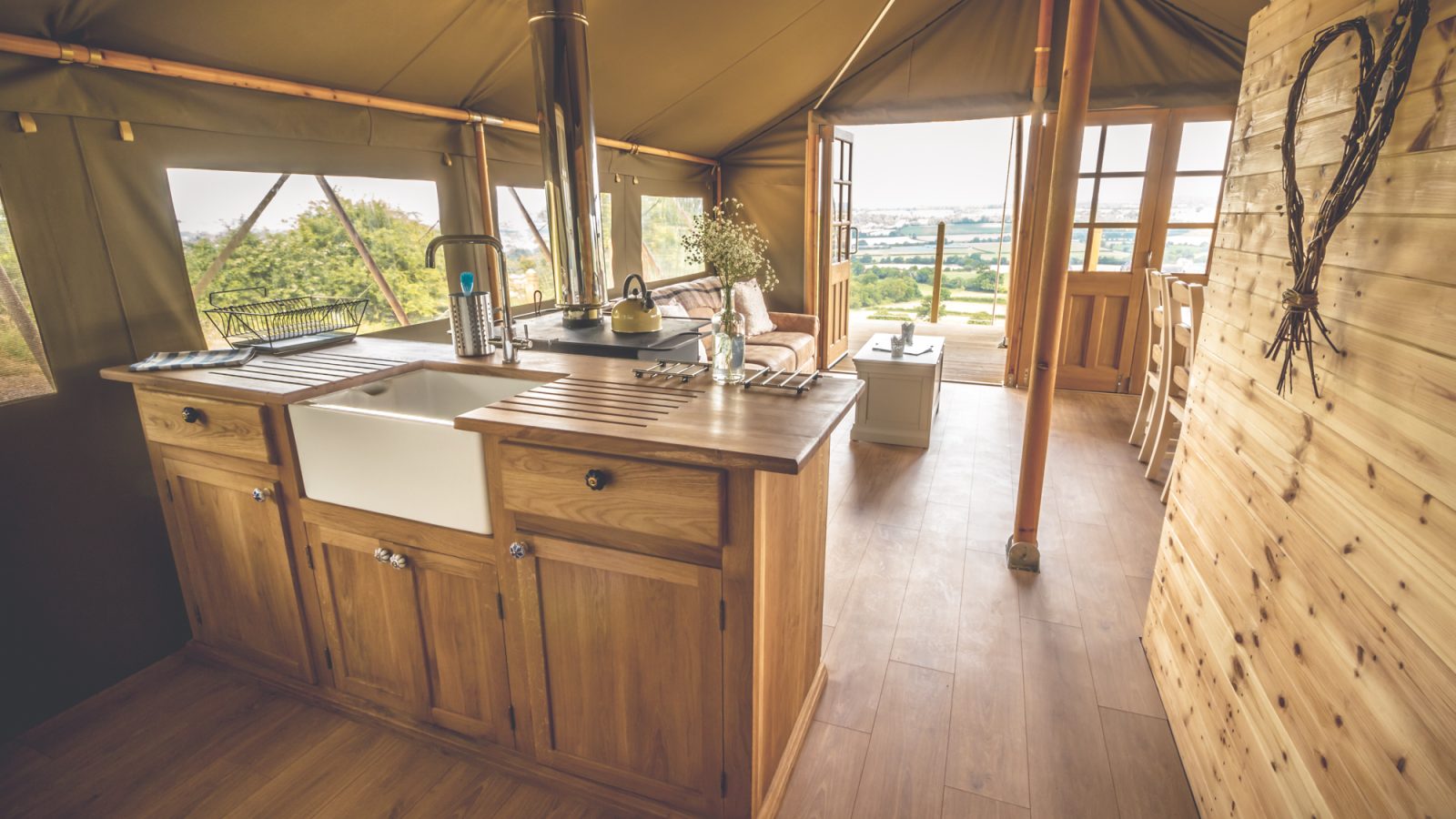 A rustic safari tent with a cozy wooden interior featuring a kitchen area with a farmhouse sink, wooden countertops, and a stove. Large windows provide a view of outdoor greenery. A dining area with a table and chairs is visible in the background.