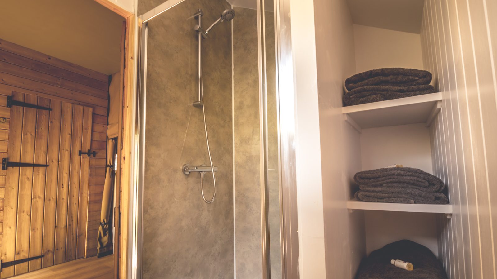 A modern bathroom featuring a glass-enclosed shower with a silver showerhead evokes the essence of a luxurious safari tent. To the right, white shelves are stocked with neatly folded dark towels and toiletries. An adjacent wooden door leads into a sauna with wooden interior walls.