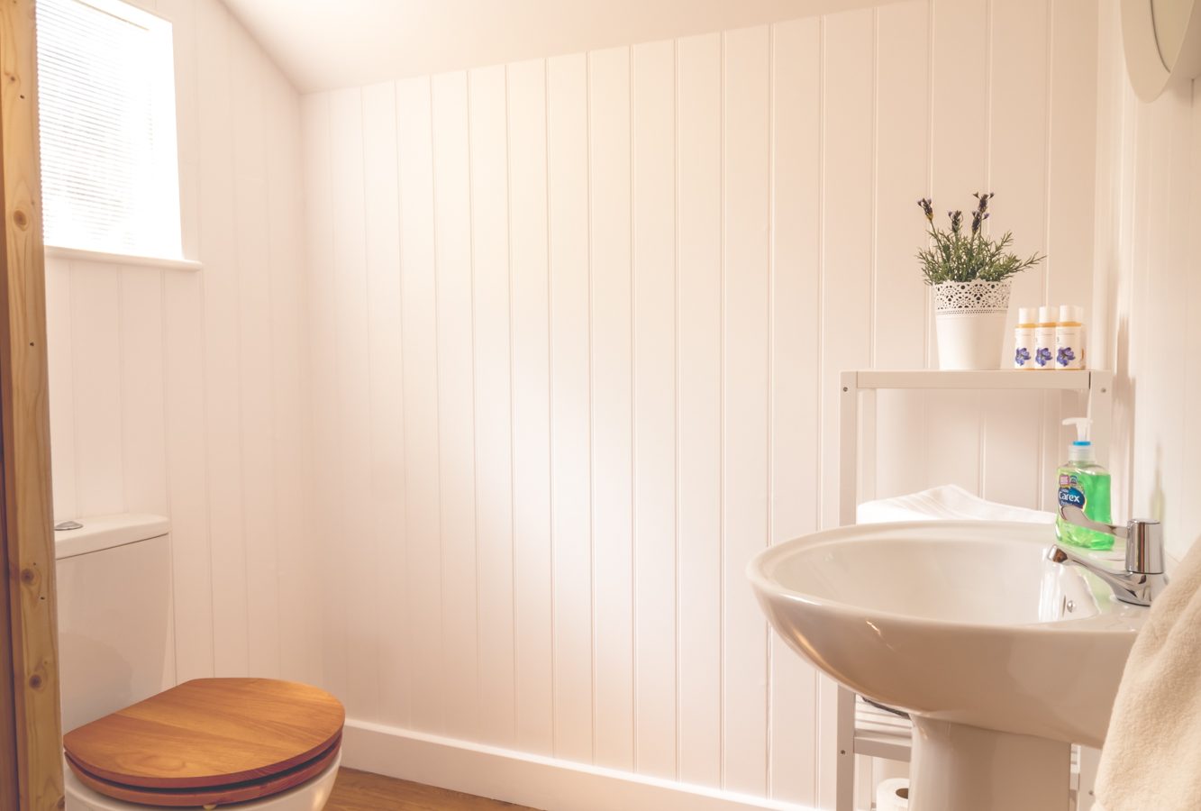 A small bathroom with white wood-paneled walls and a slanted ceiling, reminiscent of a cozy safari tent. The room features a white sink and toilet with a wooden seat, a round mirror above the sink, and a shelf with toiletries and a potted plant. The wooden floor adds warmth to the space.