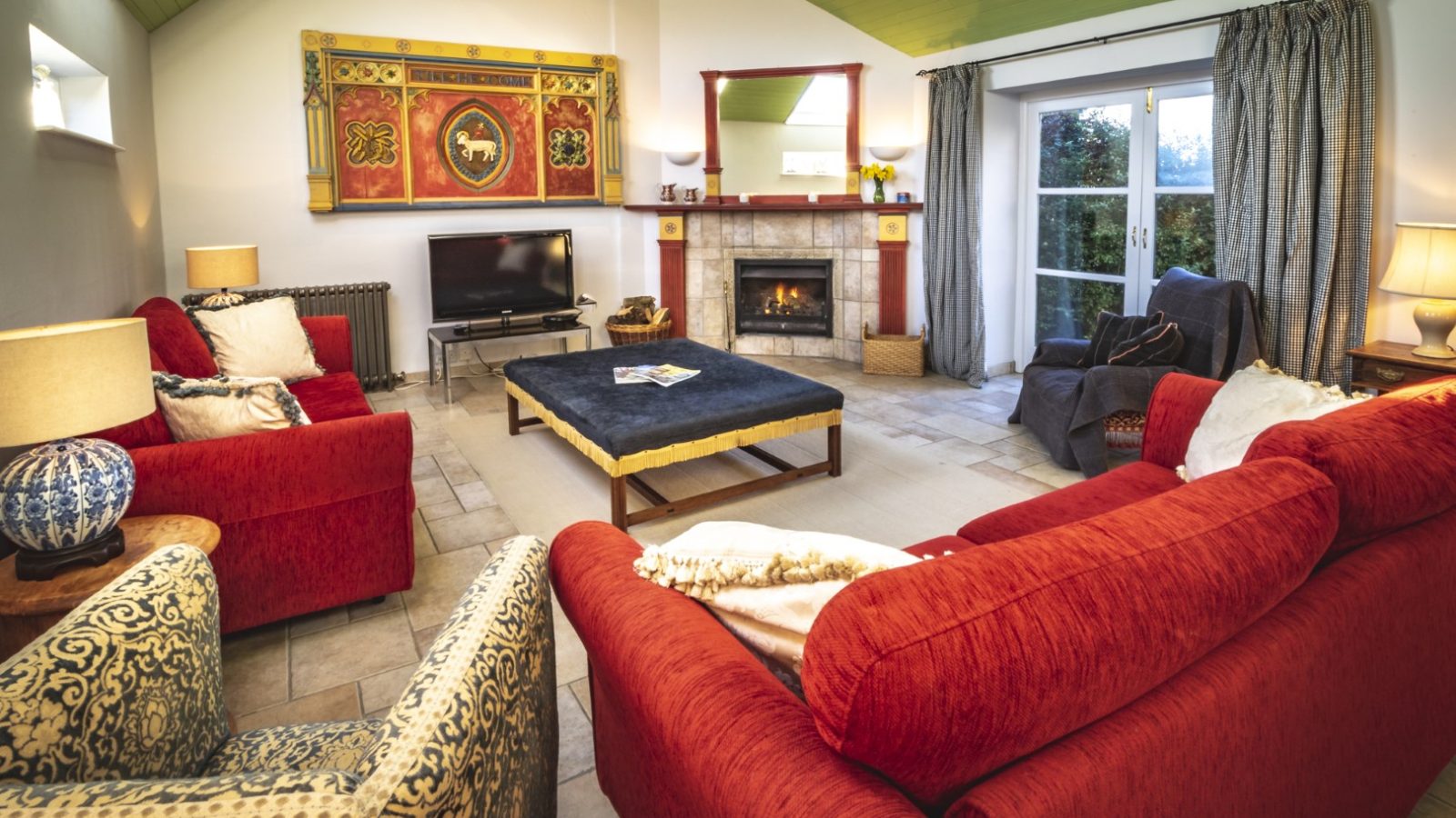 A cozy living room at Pennard Farm Cottages features two red sofas, patterned armchairs, a central dark coffee table, and a lit fireplace above a TV. Large windows with curtains let in natural light, and the walls are adorned with artwork. The ceiling is painted green.