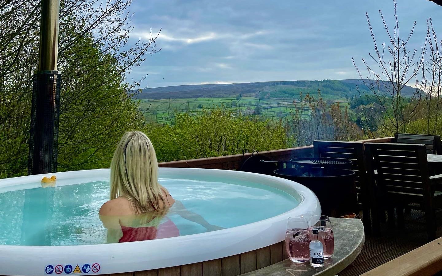 A person with blonde hair, wearing a red swimsuit, relaxes in a hot tub on the wooden deck of Howgill Lodge, overlooking a scenic valley with hills and forests. Nearby, there's a glass table with drinks and chairs, and a barbecue grill on the side.