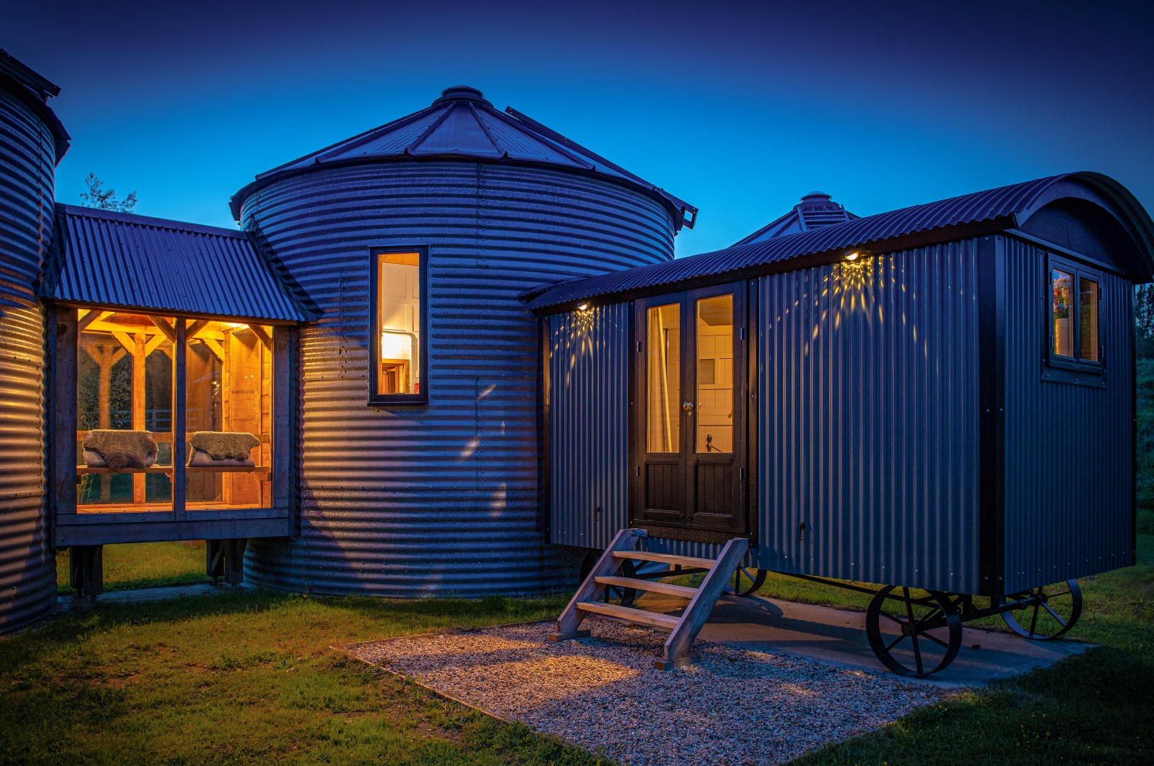 A nighttime view of a cosy, rustic retreat in Rutland, UK, featuring a small house trailer with corrugated metal siding and steps leading to the door. This coolest lodge is connected to silo-shaped structures and a covered porch with chairs, all softly illuminated against a deep blue sky.