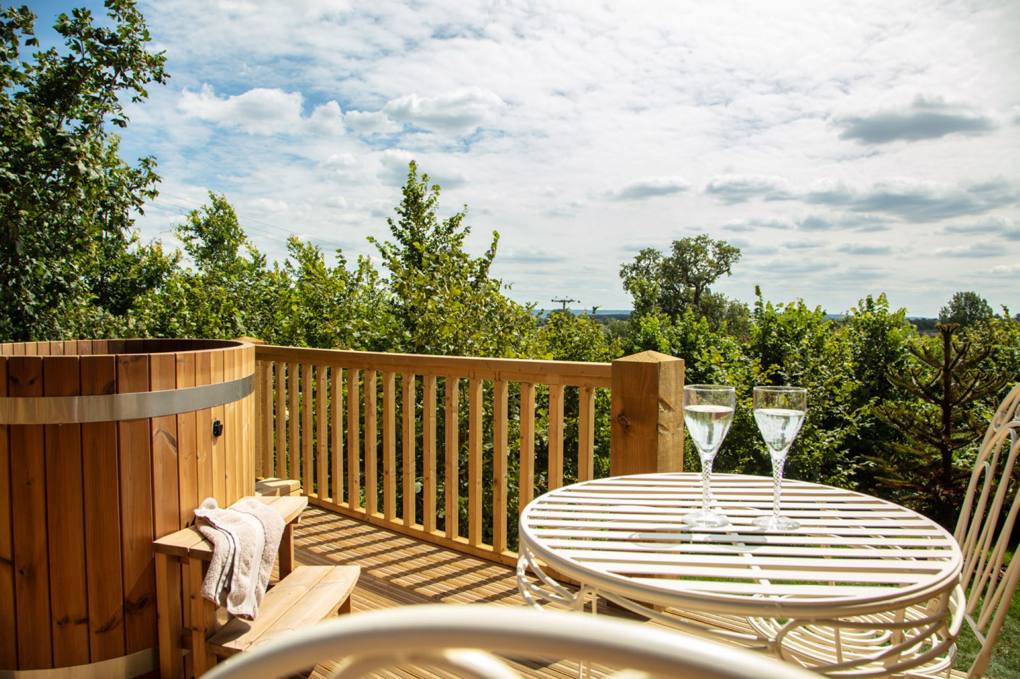 A serene outdoor patio, features a wooden hot tub and a small round table with two glasses of white wine. The patio is surrounded by lush greenery and trees under a partly cloudy sky, creating a peaceful, relaxing atmosphere. Idyllic romantic accommodation near London