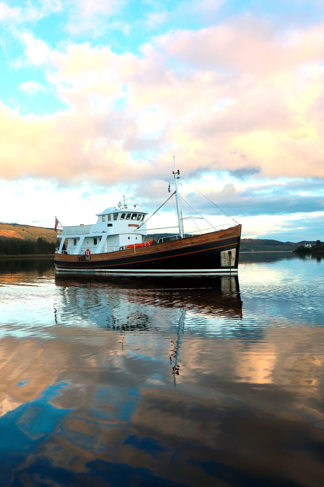 A boat peacefully floats on a calm lake with reflections of the colorful sky and clouds on the water's surface. The background shows distant, tree-lined shores and softly illuminated hills under a dramatic, partly cloudy sky.