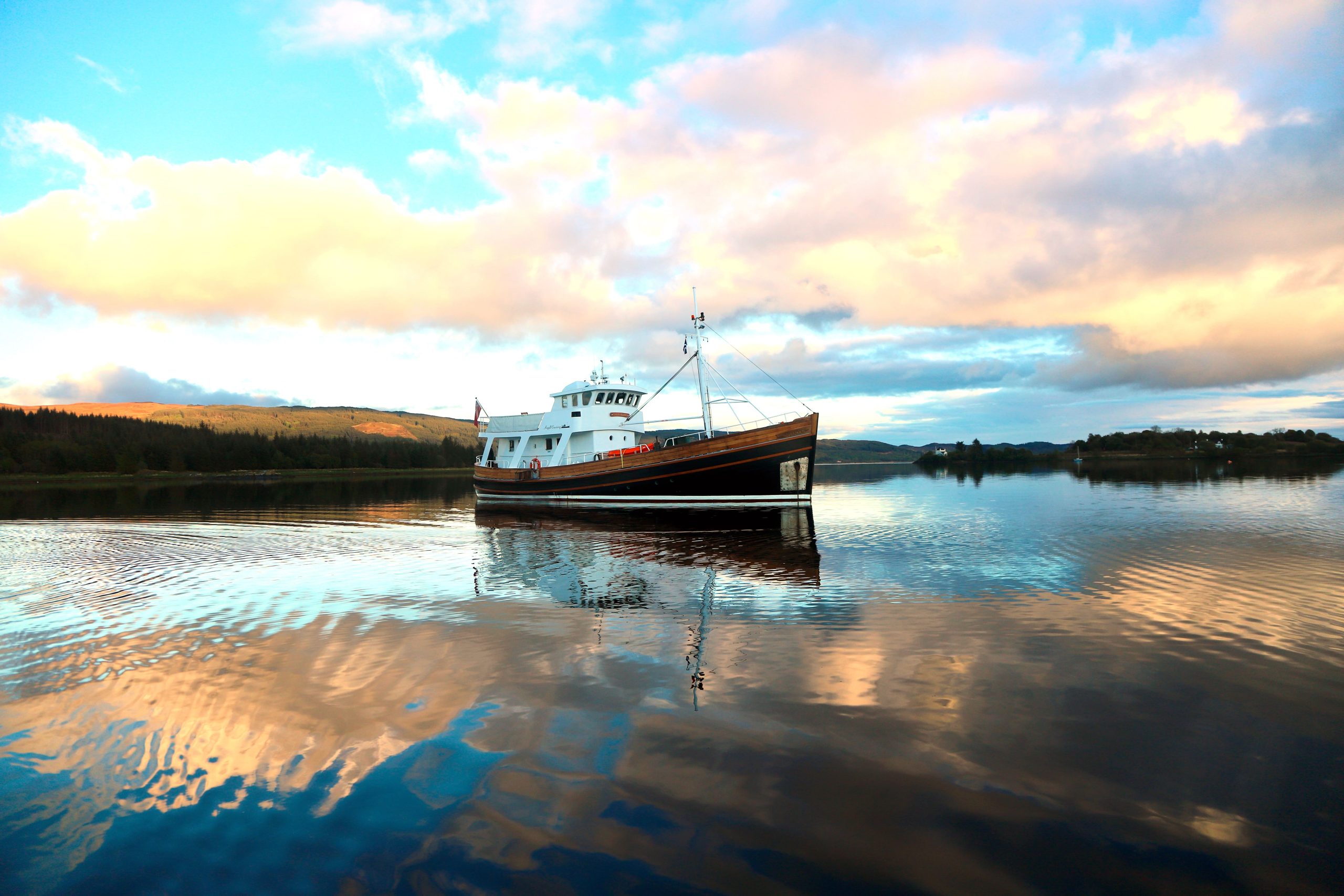 A boat peacefully floats on a calm lake with reflections of the colorful sky and clouds on the water's surface. The background shows distant, tree-lined shores and softly illuminated hills under a dramatic, partly cloudy sky.