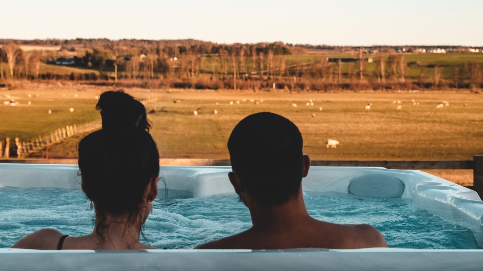 Two people relax in a hot tub at Bachilton, overlooking a scenic countryside with rolling fields and grazing sheep in the distance. The sun is setting, casting a warm glow over this idyllic farm landscape, making it the perfect spot for a tranquil holiday.