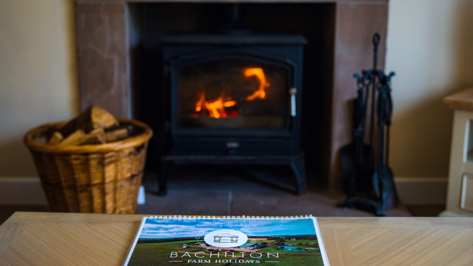 A cozy scene featuring a lit fireplace with a burning fire in a living room. In front of it is a coffee table with a brochure for Bachilton Farm Holidays. To the left of the fireplace, there is a wicker basket filled with firewood. Fireplace tools are on the right.