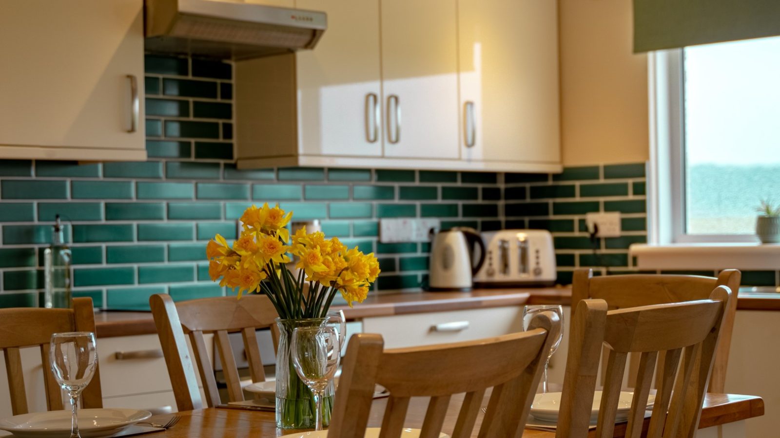 A well-lit kitchen at Bachilton Farm features white cabinets and a green subway tile backsplash. A wooden dining table adorned with a vase of yellow daffodils is set with plates, glasses, and cutlery. Appliances including a kettle and toaster sit on the wooden countertops, perfect for cozy farm holidays.