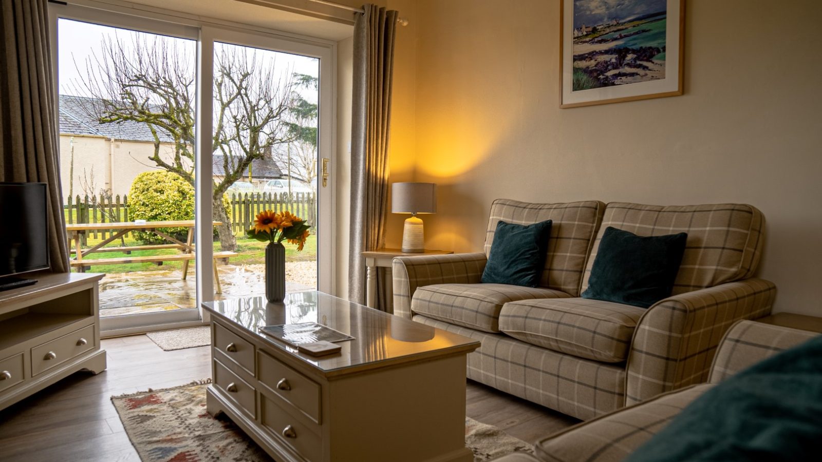 A cozy living room with a plaid sofa and armchair, two dark green cushions, a white coffee table with sunflowers in a vase, a table lamp, a framed landscape painting from Bachilton Farm, and glass doors leading to a garden with a leafless tree and wooden fence perfect for the holidays.