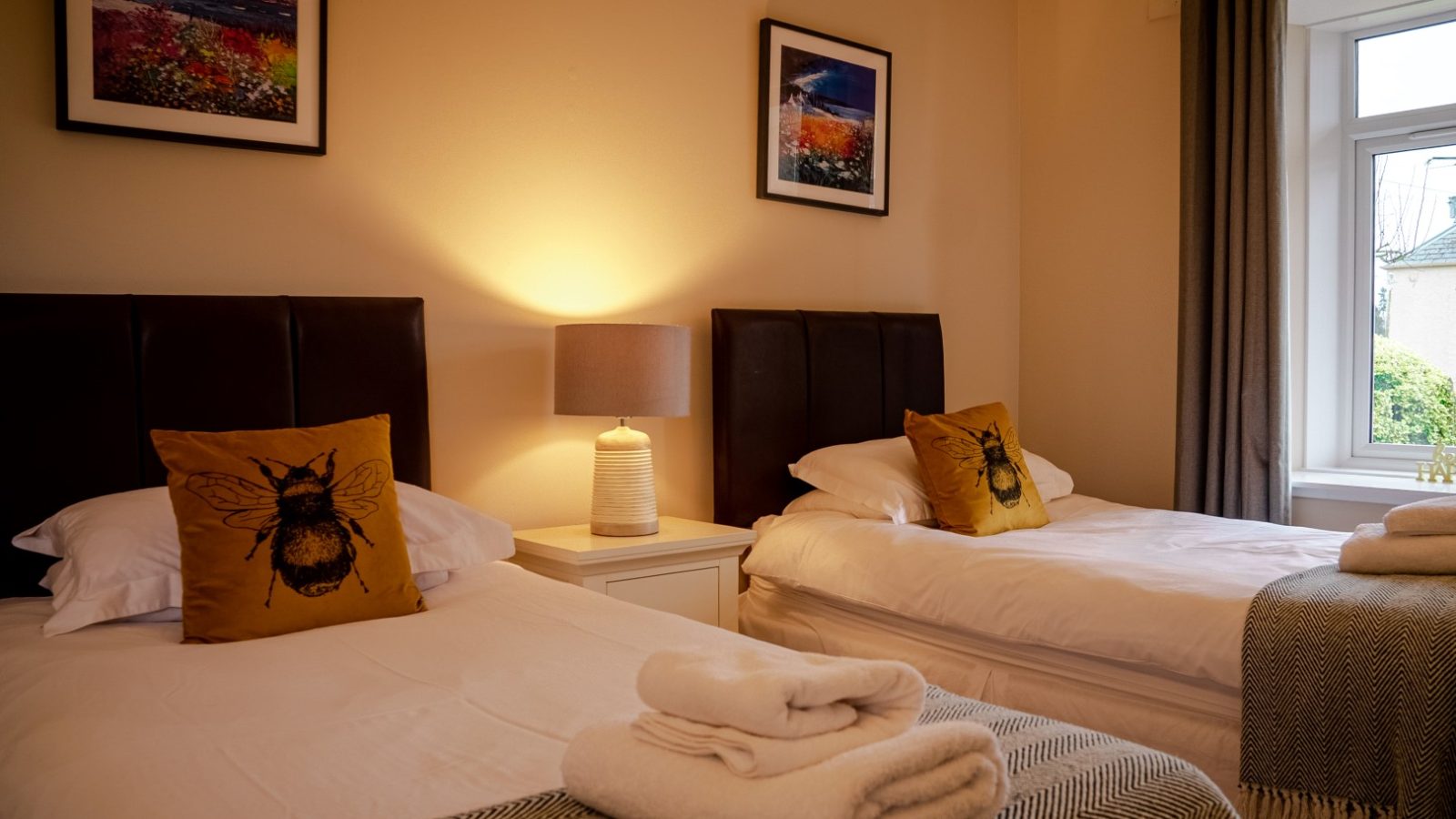 A cozy bedroom at Bachilton Farm Holidays features two single beds, each with a black headboard. Both beds have white linens, bee-themed pillows, and rolled white towels at the foot. A lit table lamp sits on a nightstand between the beds. The walls boast framed landscape artwork, and there's a window with closed curtains.