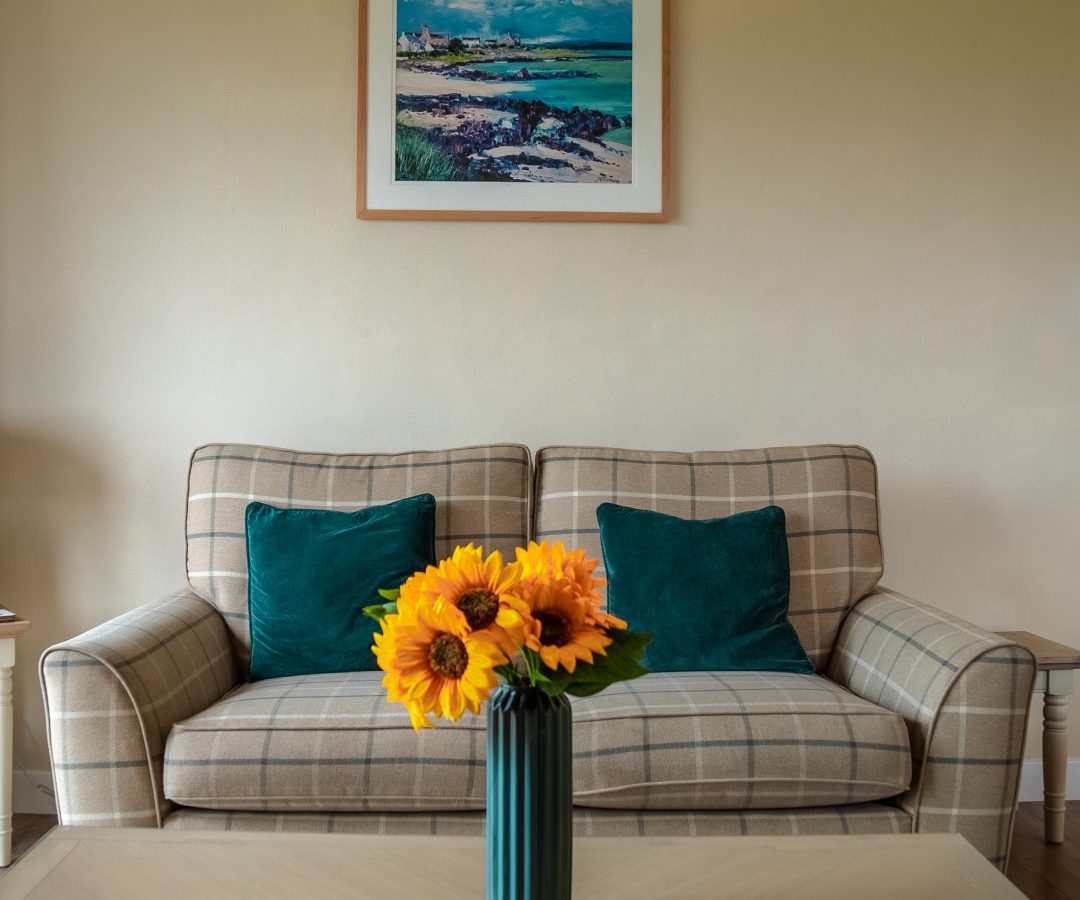 A cozy living room with a plaid loveseat adorned with two green cushions. A blue ceramic vase holding bright yellow sunflowers sits on a wooden coffee table in the foreground. Above the loveseat hangs a framed painting of a coastal landscape, evoking memories of Bachilton farm holidays.