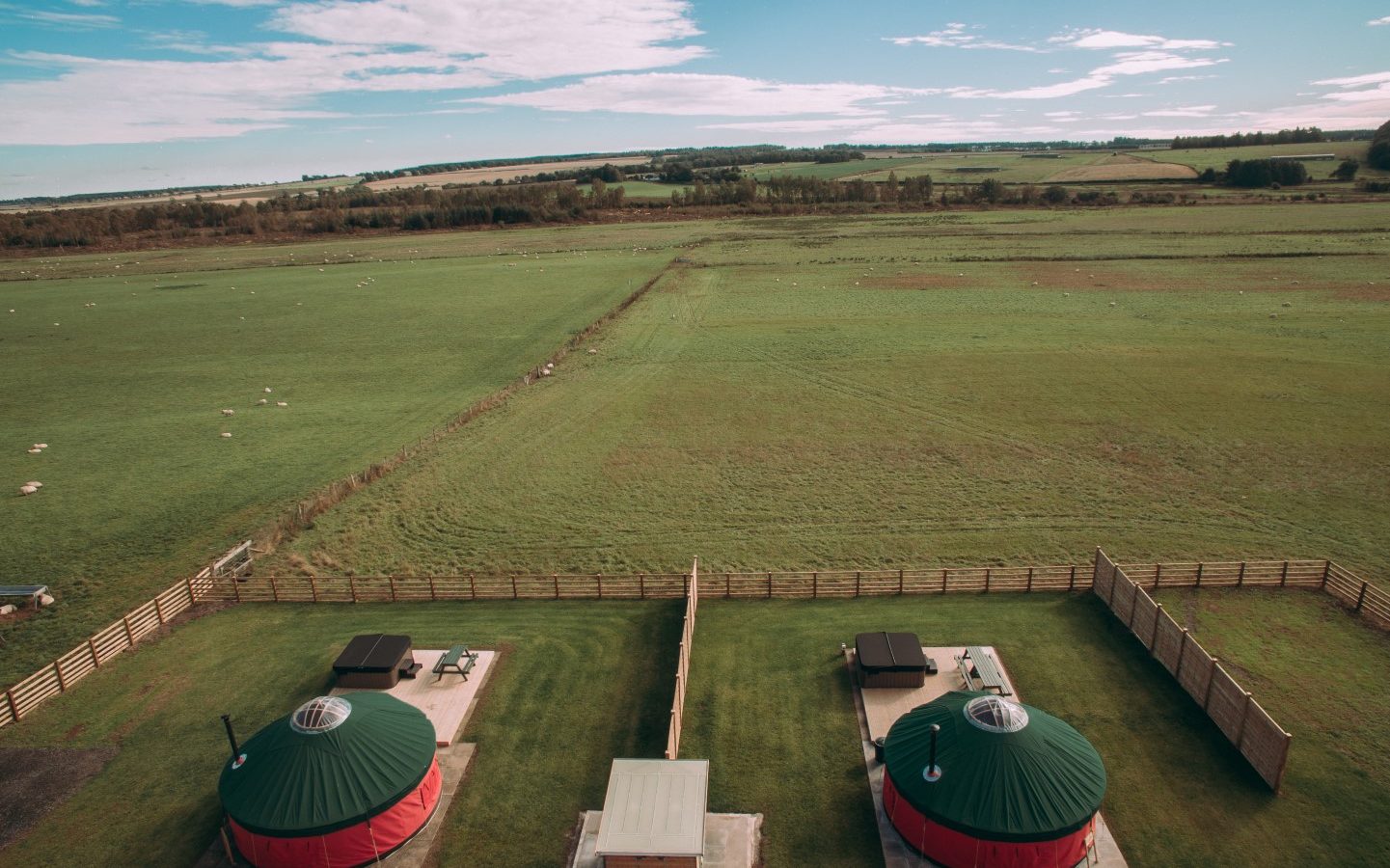 Aerial view of two yurts with green roofs and red walls at Bachilton Farm Holidays. Each yurt boasts a deck with outdoor seating, nestled in fenced grassy areas. The surrounding fields stretch into the distance under a blue sky with scattered clouds, offering an idyllic retreat.