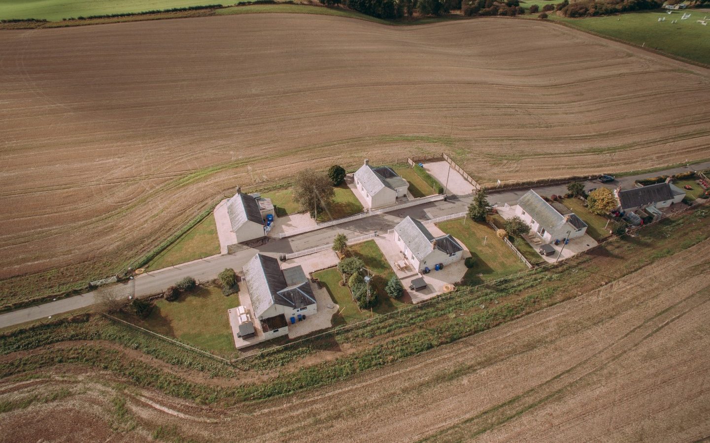 An aerial view of a small rural community consisting of a few houses lined along a single road. The homes are surrounded by vast, plowed fields and some green patches, bordered by trees in the background. This idyllic landscape is reminiscent of Bachilton Farm Holidays, with its primarily agricultural charm.