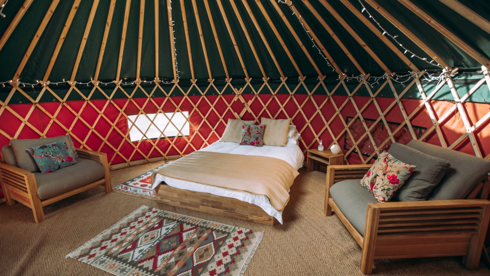 The interior of a cozy Bachilton yurt features a neatly made bed, two wooden armchairs with colorful cushions, a small side table, and a geometric-patterned rug. The walls are lined with lattice, and string lights hang from the ceiling, perfect for an idyllic farm holiday retreat.