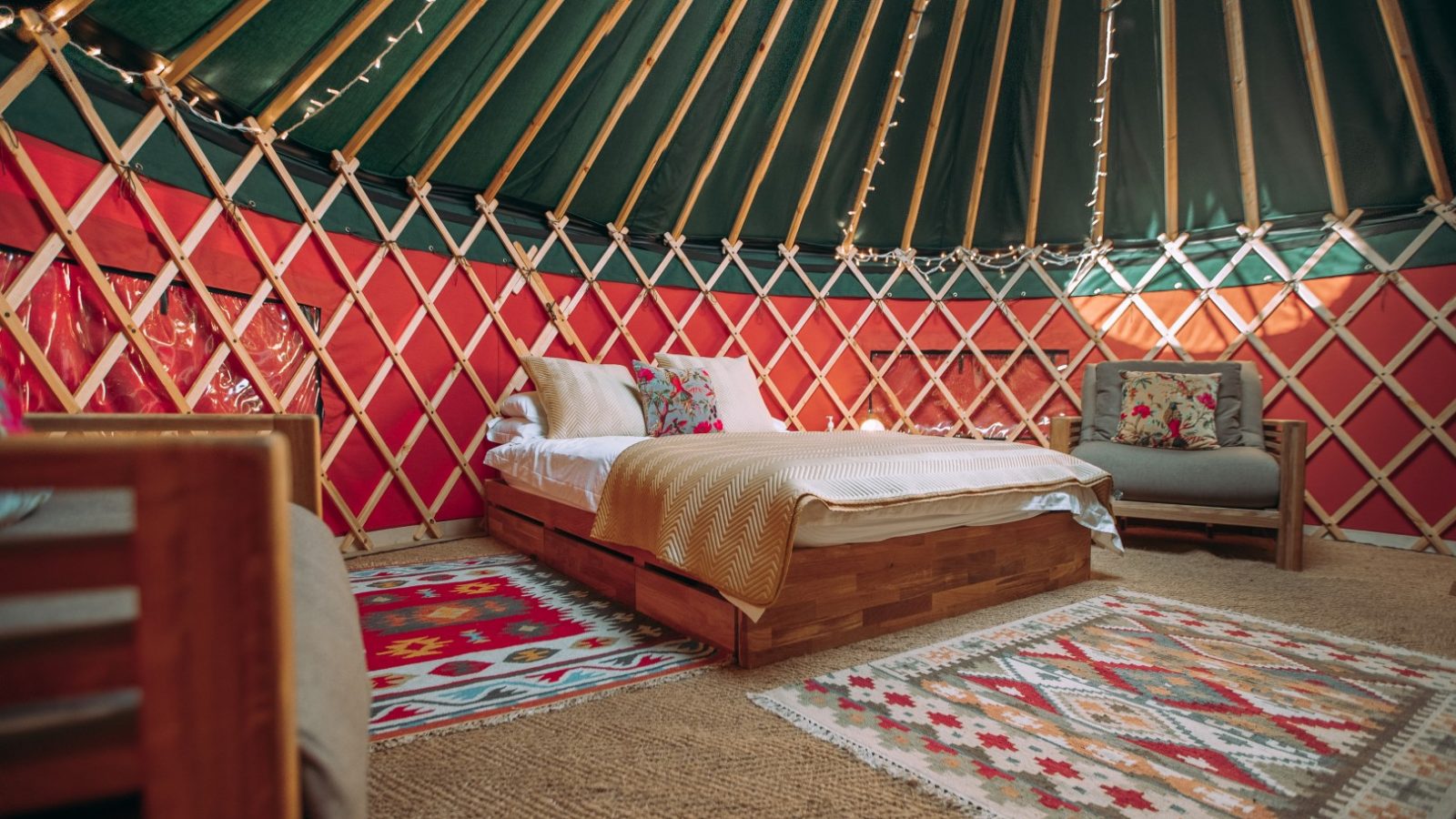 An interior view of a cozy yurt at Bachilton Farm, featuring a wooden bed with white linens and colorful pillows. The walls are adorned with a lattice framework and bright red fabric. String lights hang around the ceiling, and various patterned rugs cover the floor, adding warmth to your holiday retreat.