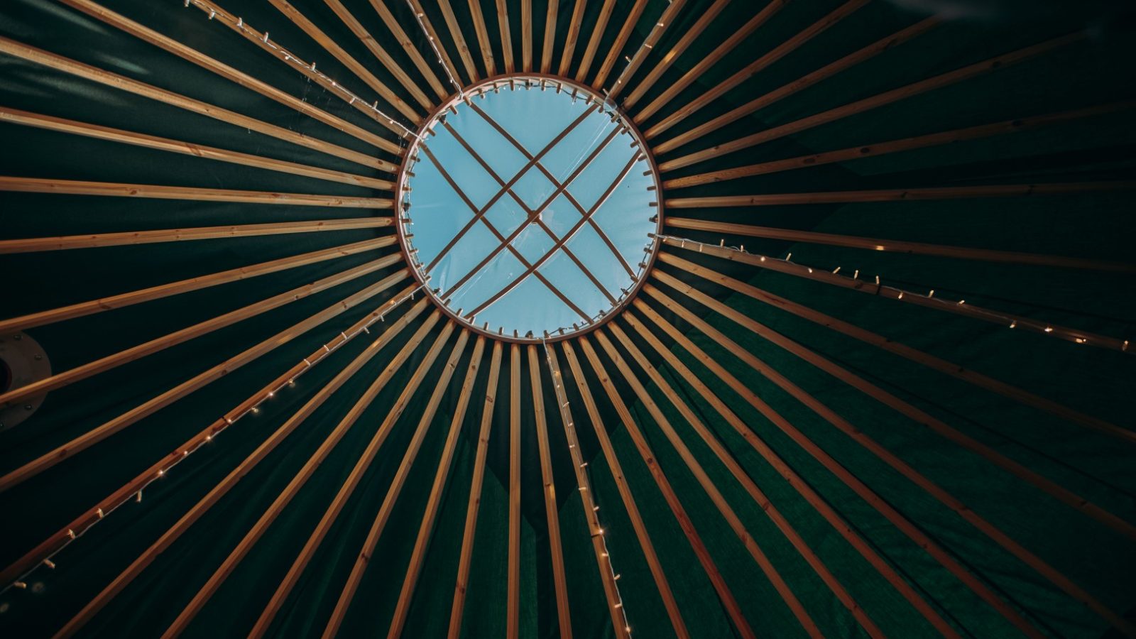 A view of a yurt's roof interior at Bachilton Farm Holidays, showcasing the circular skylight and wooden roof supporting beams radiating outward in a spoke pattern. The blue sky is visible through the skylight, contrasting with the dark green interior fabric of the yurt.