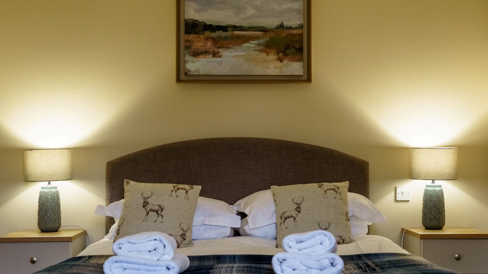 A cozy bedroom in Bachilton with a double bed featuring a checkered blue blanket, two neatly folded towels at the foot, and two pillows with deer prints. Matching bedside tables with lamps sit on either side, and a landscape painting hangs above the bed—perfect for farm holidays.