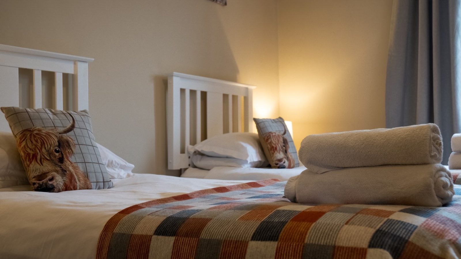 A cozy bedroom at Bachilton Farm features two single beds side by side, each with a white frame and animal-themed cushion. A colorful checkered blanket covers the foot of the beds. A stack of folded towels is placed on the closest bed, and a bedside lamp softly illuminates the room, perfect for holidays.