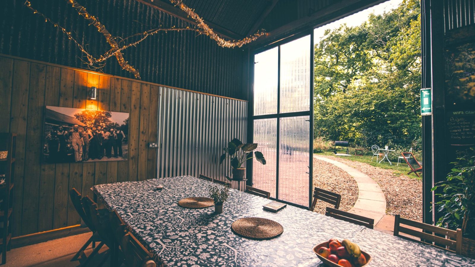 A cozy, rustic room with a long table covered in a patterned tablecloth, wooden chairs, and a bowl of fruit. Twinkling string lights adorn the wooden walls of Nantseren Cottage. Large open windows reveal a path leading to a green garden outside.