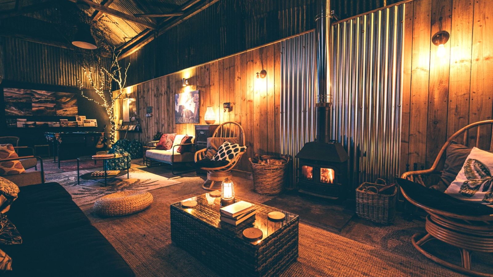 A cozy, dimly lit interior with a rustic design featuring corrugated metal walls, a wood-burning stove, and Nantseren wicker chairs. It also boasts a wooden ceiling, cushioned sofa, and central wooden coffee table adorned with a candle. Warm, ambient lighting enhances the inviting atmosphere.