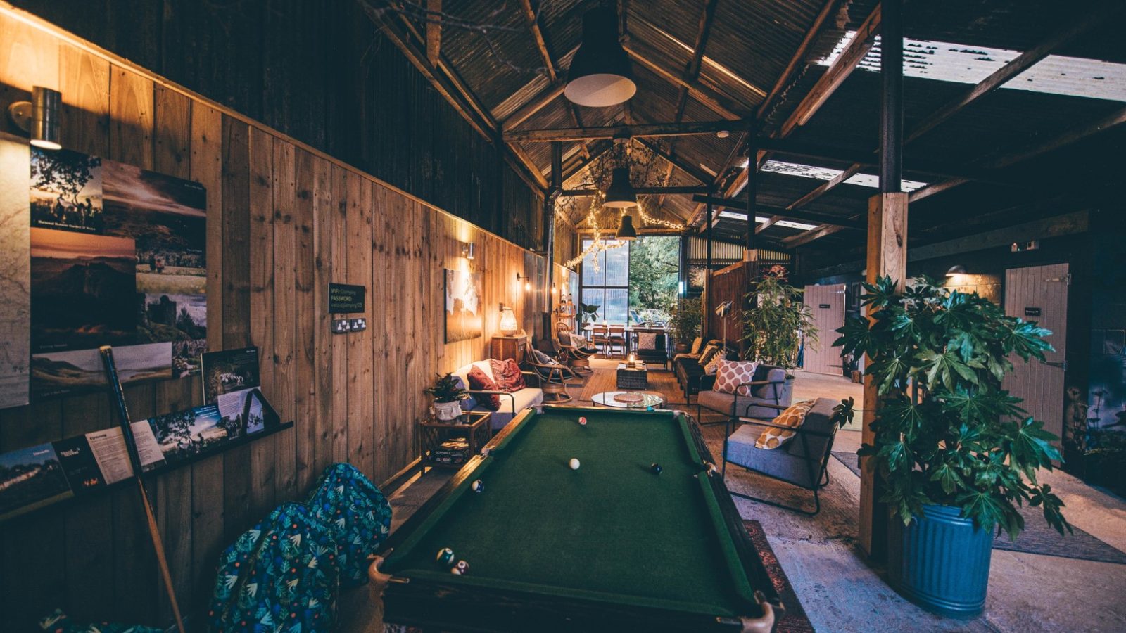 Cozy indoor space with a rustic, wooden ceiling, and a softly lit atmosphere. A green pool table by Nantseren occupies the foreground, surrounded by sofas, plants, and vintage decor. A large window at the back lets in natural light, highlighting a relaxed, inviting ambiance.