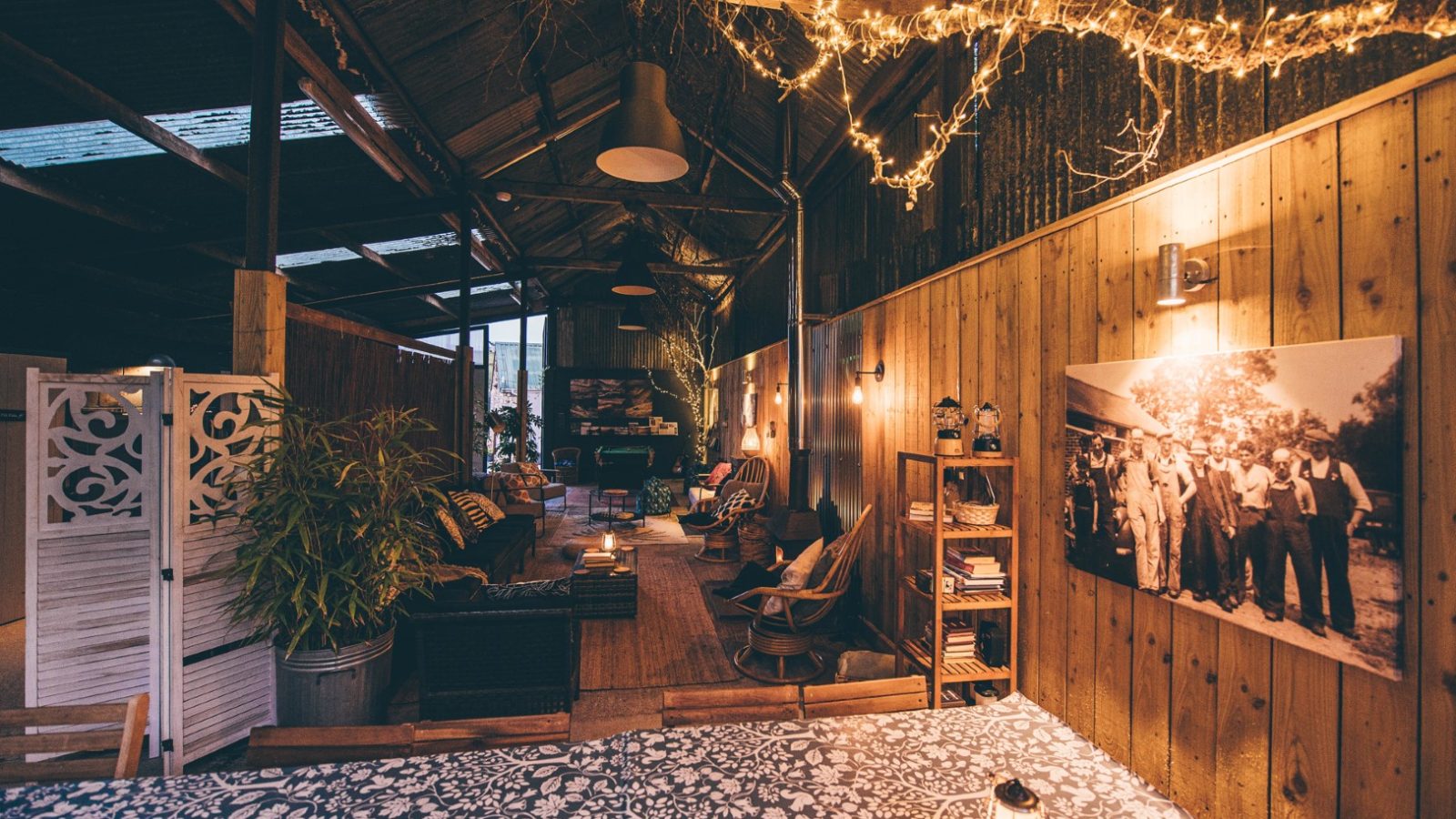 A cozy, rustic indoor space with wooden walls and a high, corrugated metal ceiling. String lights hang from the ceiling, illuminating the area. The Nantseren-inspired space includes a seating area with sofas, plants, a bookshelf, a patterned rug, and a black-and-white group photo on the wall.