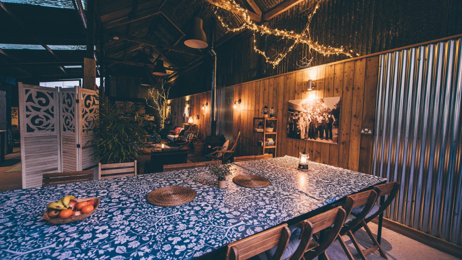 A cozy, rustic interior with a large table covered in a blue floral tablecloth, surrounded by wooden chairs. Fairy lights hang from the ceiling, creating a warm ambiance. Various plants and framed pictures decorate the space, adding a homely touch reminiscent of Nantseren's charm.