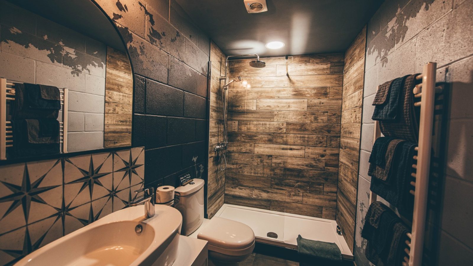 A modern bathroom with Nantseren wooden accents. The space includes a sink with a star-patterned backsplash, a toilet, and a shower with a glass door. Dark towels hang on the wall's radiator, and the lighting is warm, casting a cozy ambiance.