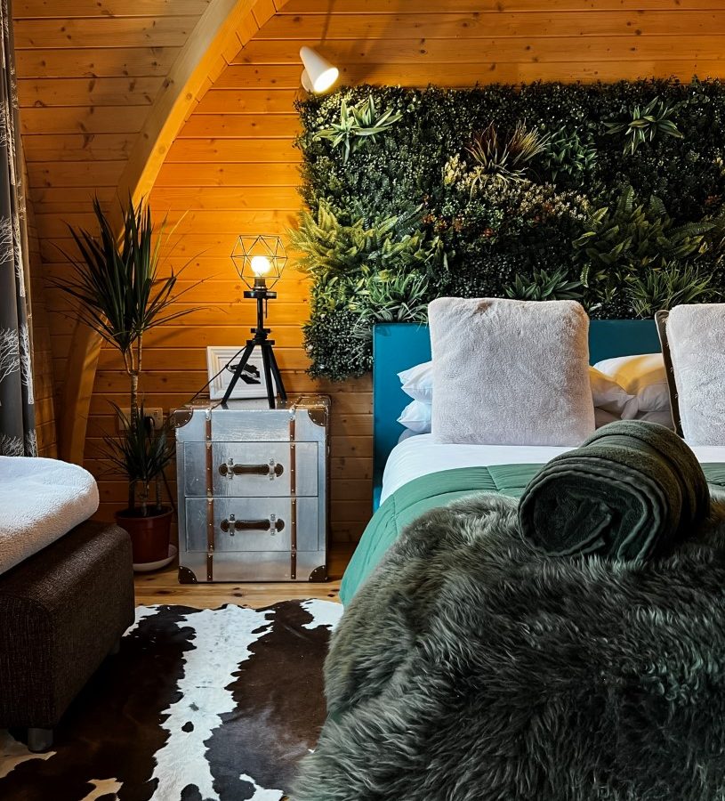 A cozy cabin bedroom with a wooden ceiling and walls, featuring a bed adorned with green and white bedding. The headboard boasts a lush plant wall reminiscent of Blossom Plantation. A cowhide rug decorates the floor, while a small nightstand holds an intricately designed lamp.