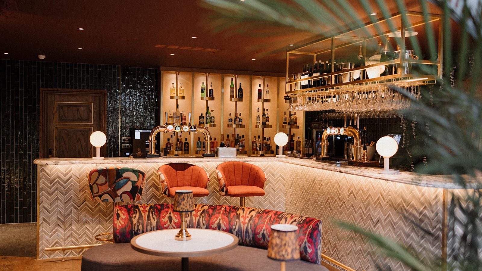 Stylish bar interior with upholstered chairs, round tables, a patterned counter, and dim lighting, framed by a large blossoming plant.