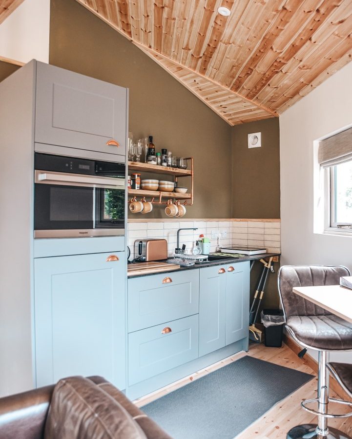 A compact kitchen with light blue cabinets, a wooden countertop, and a sleek oven. Shelves hold jars and mugs. The room has a wood-paneled ceiling and light brown flooring. A window provides natural light, overlooking the serene view of Blossom Plantation, with a small dining area visible.