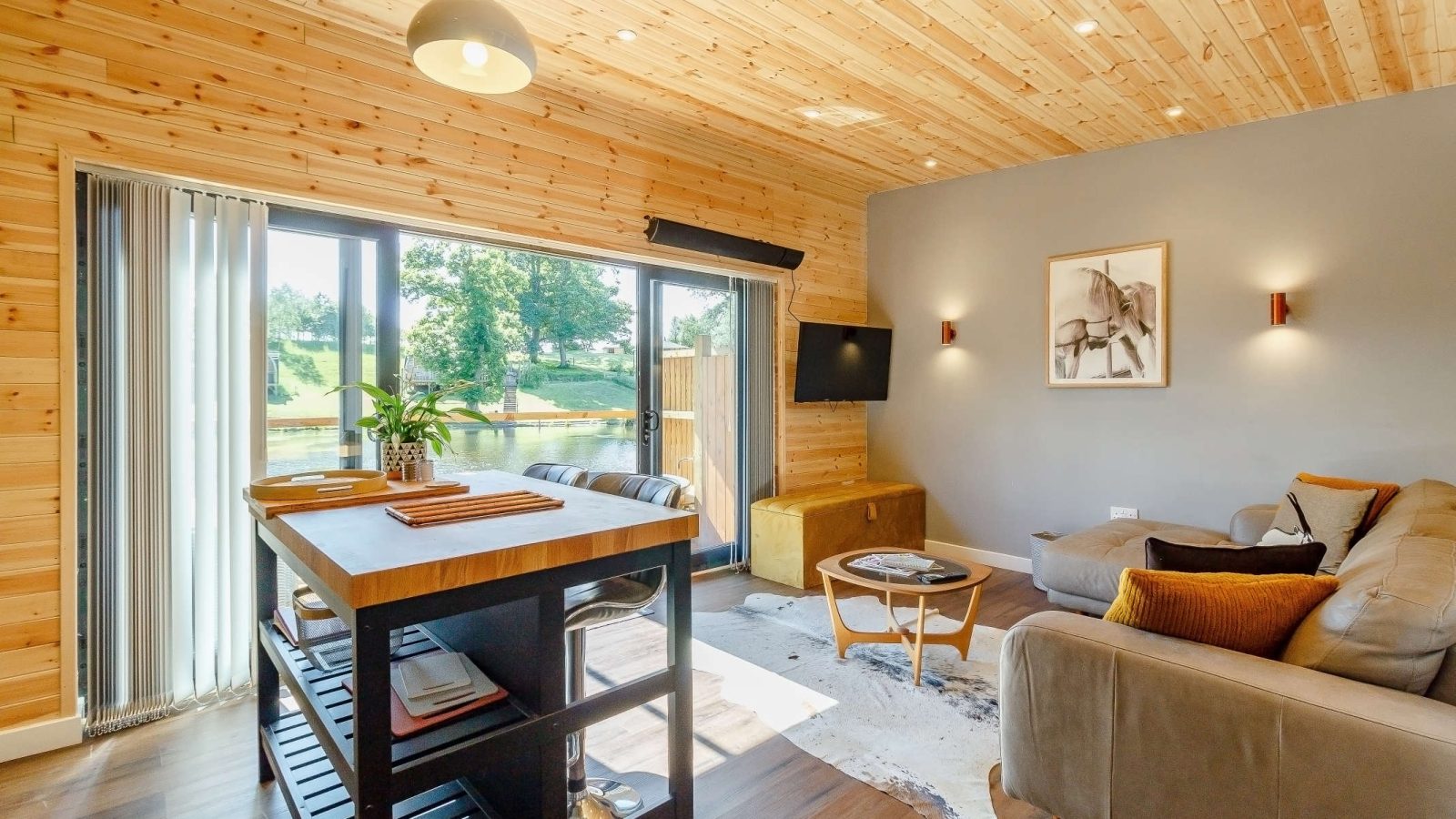 A cozy living room with wooden walls and ceiling, featuring a large window that frames the blossoming trees outside. There's a sofa, a wooden coffee table on a rug, a wall-mounted TV, and a high table with stools in the foreground. Warm lighting enhances the space.