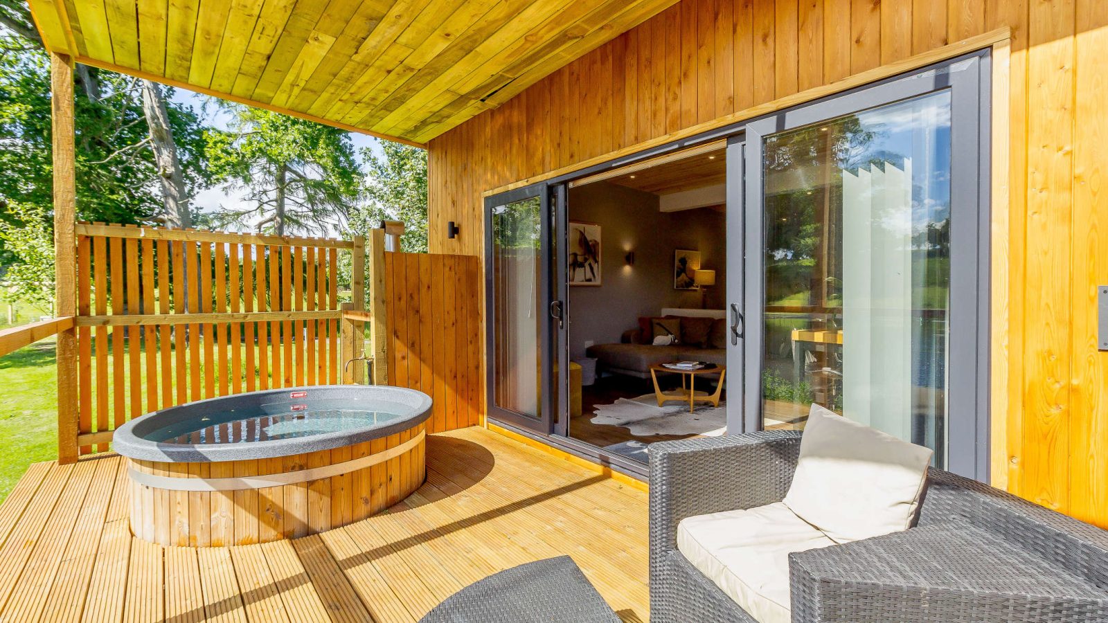 A wooden deck with a hot tub surrounded by a wooden privacy screen offers serene relaxation amidst nature. Sliding glass doors lead to a cozy interior with a couch and chairs. Sunlight dances on the deck, overlooking the lush trees of Blossom Plantation.