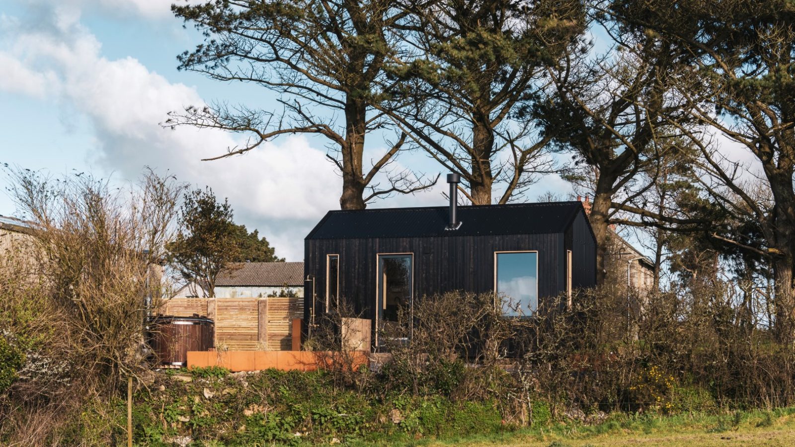 A small, modern black house nestled among tall trees and shrubs in a rural area on a sunny day. The house, reminiscent of a lookout, features large windows and minimalist design, with clear blue skies and light clouds in the background. An open grassy field at Bogee Farm is seen in front of the house.