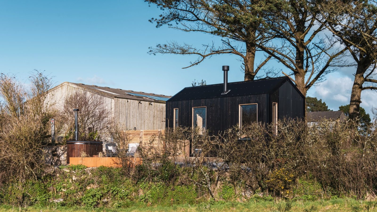 A small black wooden cabin with a chimney and large windows is nestled among trees and bushes in the rural expanse of Bogee Farm. A rustic barn stands behind the cabin, known locally as The Lookout. The scene is set against a clear blue sky.