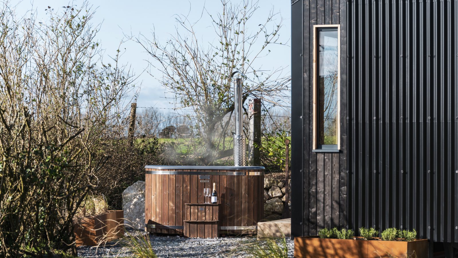 A modern black tiny house, known as The Lookout, stands beside a wooden hot tub nestled in a garden with gravel and raised planters. Leafless shrubs and trees surround the area on a clear day, with steam rising from the hot tub, adding a touch of coziness to the serene outdoor setting at Bogee Farm.