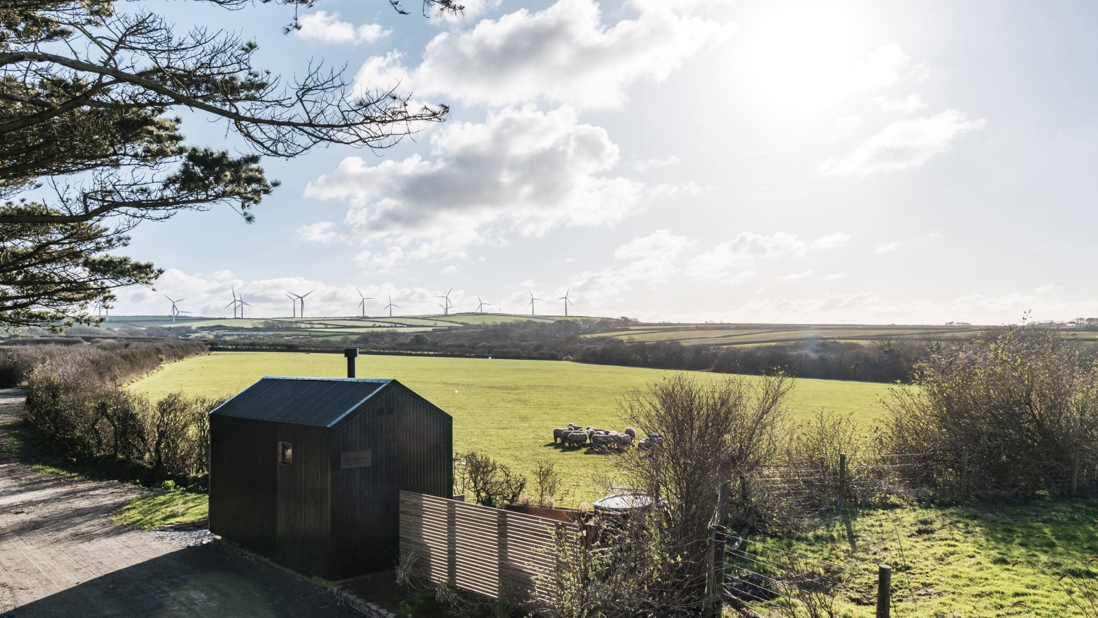 A sunlit rural landscape featuring a small wooden cabin with a green roof in the foreground. Beyond the cozy cabin, perched like a lookout at Bogee Farm, a vast green field stretches out to the horizon, dotted with wind turbines. Overhead, the sky is partly cloudy with the sun shining brightly.