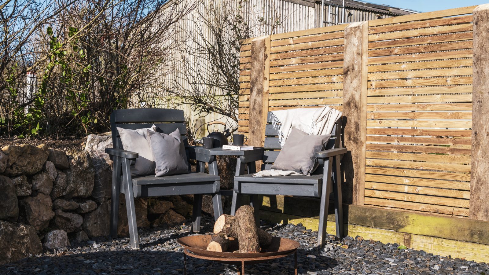 A cozy outdoor seating area at Bogee Farm with two gray wooden chairs, each with gray cushions, facing a small round fire pit containing firewood. The lookout area is surrounded by a wooden privacy screen and stone walls, under a clear blue sky.