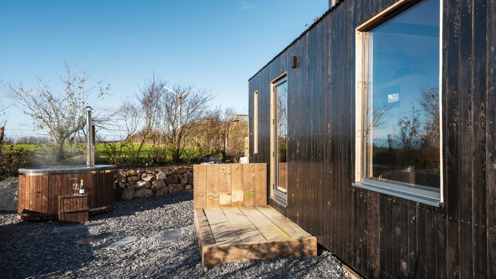 A modern black wooden cabin with large windows is situated outdoors on the gravel surface of Bogee Farm. A step made of wooden planks leads to the entrance, and to the left, a circular hot tub with wooden surroundings catches the eye. Sparse trees and a stone wall frame this serene lookout.