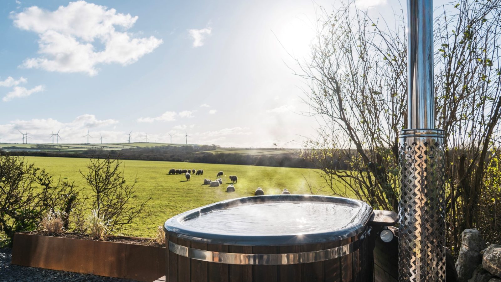 A hot tub at The Lookout sits outdoors, overlooking a vast green field with grazing sheep and cows. Wind turbines are visible in the distance against a blue sky with scattered clouds. Bushes and branches surround the hot tub area at Bogee Farm, creating a serene sanctuary.