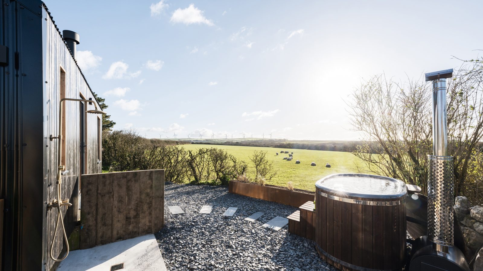 A serene outdoor scene at Bogee Farm features a small deck with a wooden hot tub and an outdoor shower. The area is surrounded by gravel and overlooks a green field dotted with hay bales under a bright, sunny sky. Sparse trees frame the view, enhancing the sense of peaceful seclusion at The Lookout.