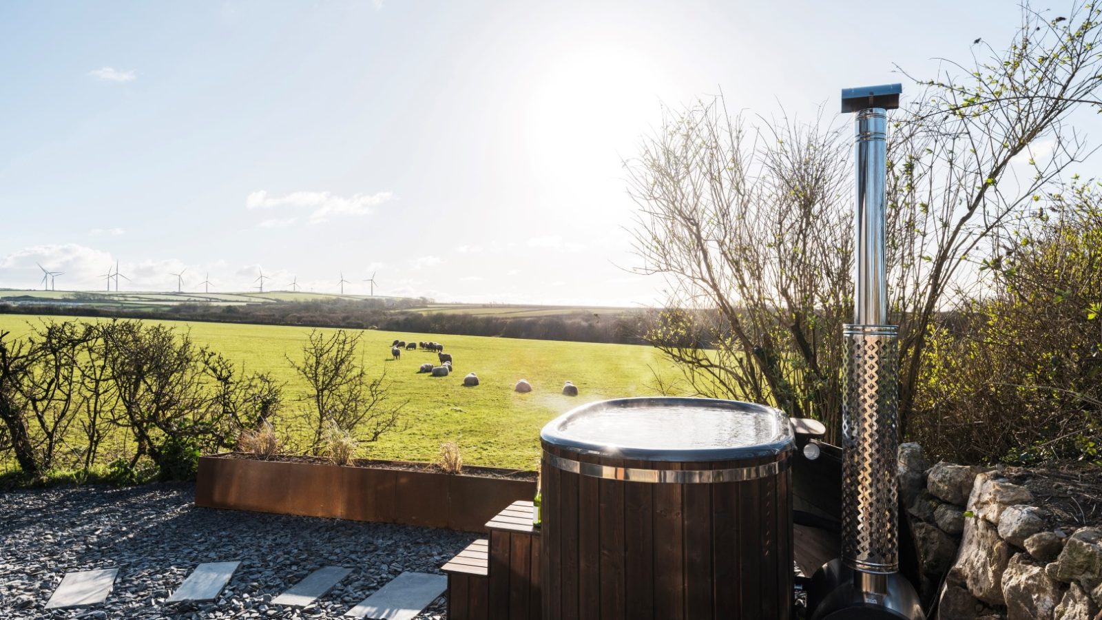 A wooden hot tub with a metal chimney is situated on a gravel patio overlooking the lush green field of Bogee Farm. The field is dotted with grazing sheep and wind turbines in the distance. Bushes and a stone wall border this idyllic lookout. The sky is clear with the sun shining brightly.