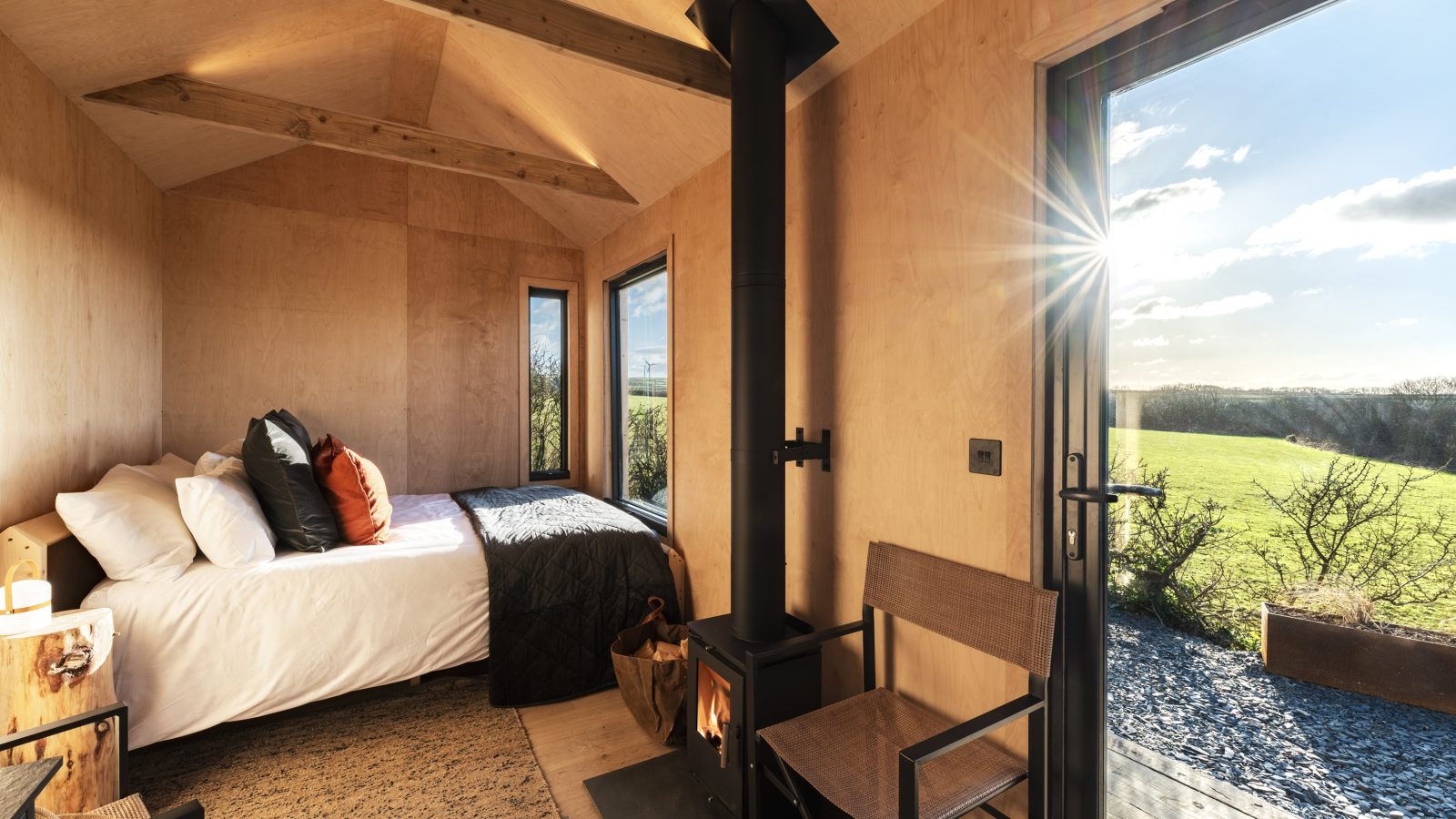 A cozy wooden cabin interior at Bogee Farm features a bed, wood-burning stove, and two chairs. Large windows and an open door reveal a grassy landscape and a clear blue sky with the sun shining. The room is warmly lit by natural sunlight.