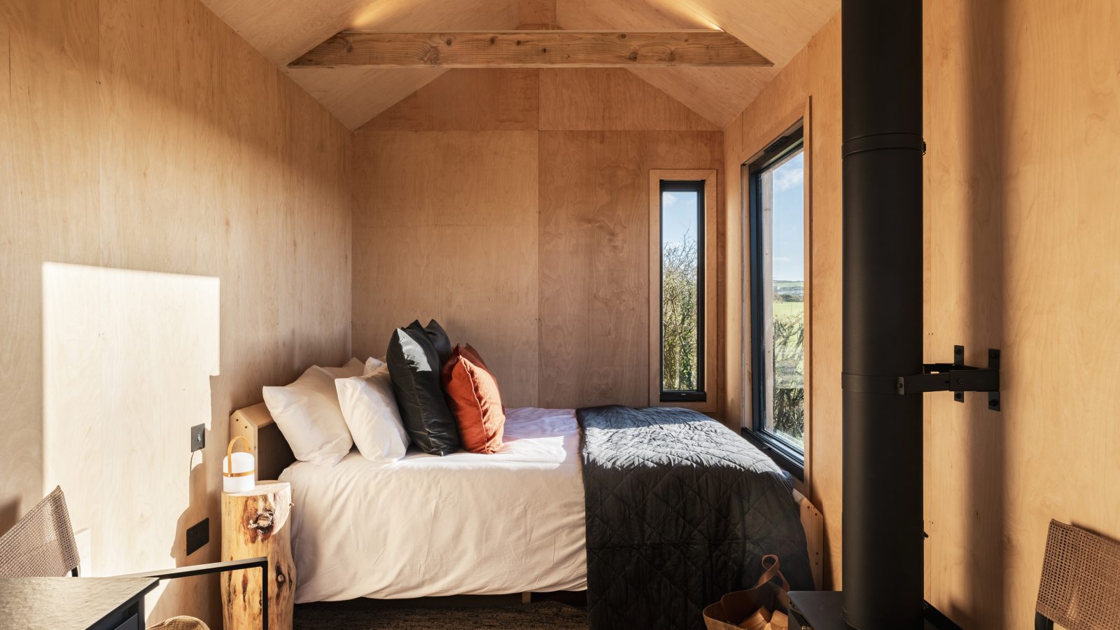 A compact, cozy bedroom at The Lookout, with natural wood walls, features a neatly made bed adorned with white linens and several pillows in varying colors. Large windows on one side let in natural light, while a small wood-burning stove sits in the corner. Rustic decor from Bogee Farm completes the space.