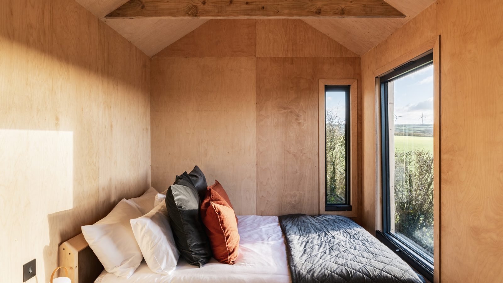 A cozy bedroom at The Lookout with wooden walls, a large bed adorned with white, black, and brown pillows, and a dark quilt. Two windows offer a view of Bogee Farm's outdoors, allowing natural light to illuminate the room. One window is large while the other is narrow and tall.